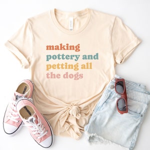 May include: A beige t-shirt with the text "making pottery and petting all the dogs" in a rainbow color scheme. The shirt is paired with light blue denim shorts and pink Converse sneakers.
