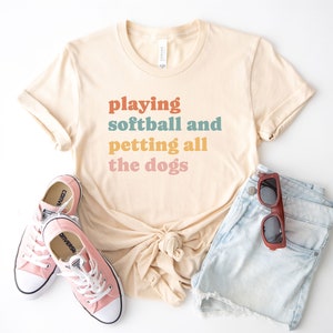 May include: A beige t-shirt with the text 'playing softball and petting all the dogs' in colorful text. The shirt is tied in a knot at the front.  A pair of pink converse shoes and a pair of blue denim shorts are also in the image.