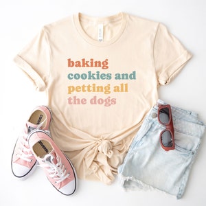 May include: A cream-colored short-sleeve t-shirt with the text "baking cookies and petting all the dogs" printed in orange, blue, yellow, and pink. The shirt is paired with light blue denim shorts, pink canvas sneakers, and dark sunglasses.