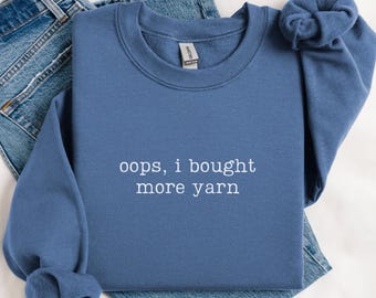May include: A blue sweatshirt with the text "oops, i bought more yarn" in white lettering. The sweatshirt is folded on top of a pair of blue jeans.