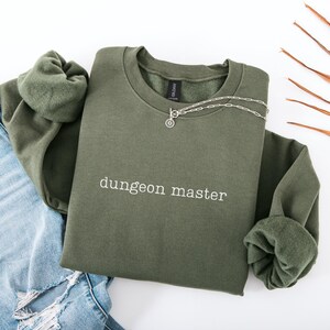 May include: An olive green sweatshirt with the text "dungeon master" in white. A silver necklace with a circular pendant is draped over the sweatshirt. The sweatshirt is folded over a pair of blue jeans.