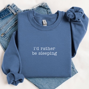 May include: A blue sweatshirt with the text "i'd rather be sleeping" in white. The sweatshirt is folded on top of a pair of blue jeans. The sweatshirt has long sleeves and a crew neck.