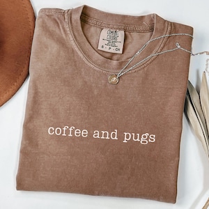 May include: A brown t-shirt with the text "coffee and pugs" in white lettering. The shirt has a distressed look and is made by Comfort Colors.