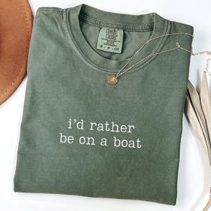 May include: An olive green short-sleeved t-shirt with a crew neckline and the white text "i'd rather be on a boat" printed on the chest. The shirt features a Comfort Colors brand label at the inner neck and is paired with a thin gold-tone necklace.