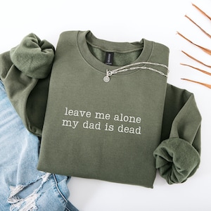 May include: An olive green sweatshirt with the text "leave me alone my dad is dead" in white. A silver necklace with a circular pendant is on the sweatshirt. The sweatshirt is folded on top of a pair of blue jeans.