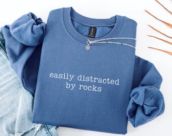 May include: A blue sweatshirt with the text "easily distracted by rocks" in white. A silver necklace with a circular pendant is draped across the sweatshirt. The sweatshirt is folded on top of a pair of blue jeans.