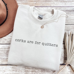 May include: A sand-colored crewneck sweatshirt featuring the black text "corks are for quitters" printed across the chest. The garment has a ribbed collar, long sleeves, and a standard fit, showcasing a simple design with clear, legible lettering.