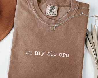 May include: A brown Comfort Colors t-shirt with the phrase "in my slp era" in white lettering. A silver necklace with a small pendant rests on the shirt. A brown hat is visible in the upper left corner.