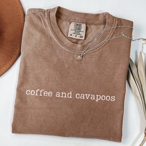 May include: A brown Comfort Colors t-shirt with the words "coffee and cavapoos" printed in white. A silver necklace with a small pendant rests on the shirt. A brown hat is visible in the upper left corner.