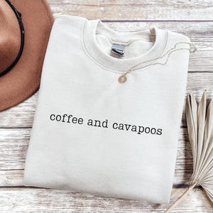 May include: A light beige sweatshirt with the text "coffee and cavapoos" in black. A gold necklace with a pendant rests on the sweatshirt. A brown hat and dried plant are in the background.