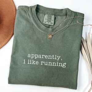 May include: An olive green t-shirt with the text "apparently, i like running" in white. A gold-colored necklace with a pendant rests on the shirt. The shirt is folded and has a tag that says "Comfort Colors".