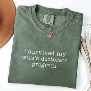 May include: Olive green Comfort Colors t-shirt with the text "i survived my wife's doctorate program" in white. A gold necklace with a pendant rests on the shirt. A brown hat and dried plant are in the background.