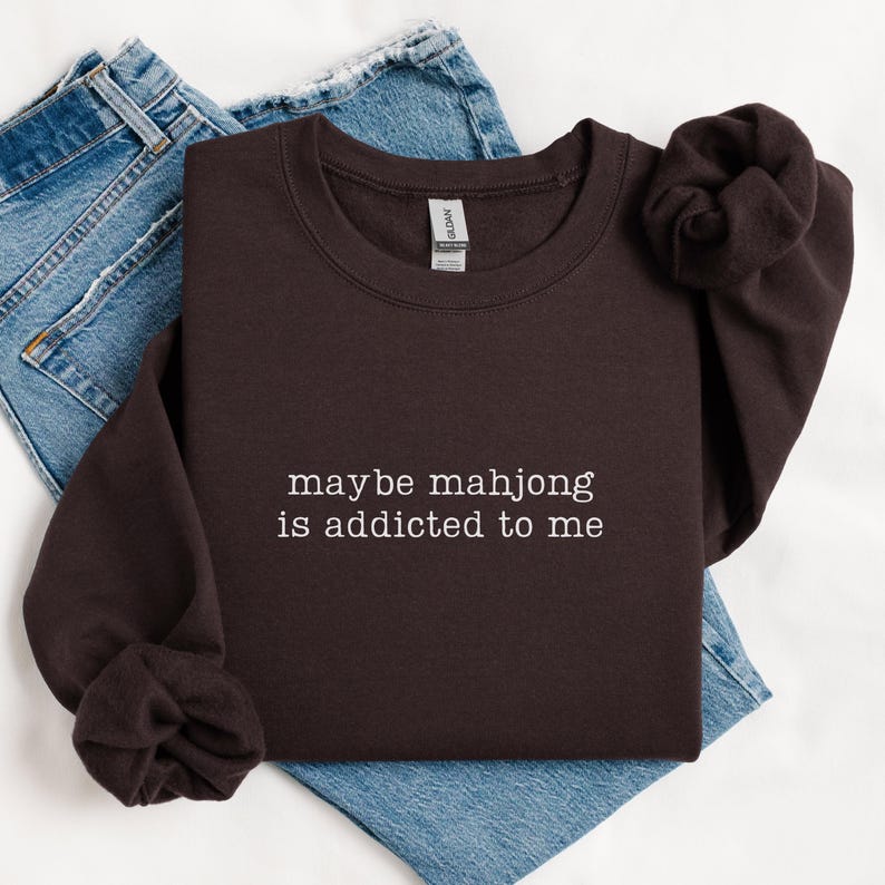 May include: A dark brown sweatshirt with the text "maybe mahjong is addicted to me" in white. The sweatshirt is folded on top of a pair of blue jeans.