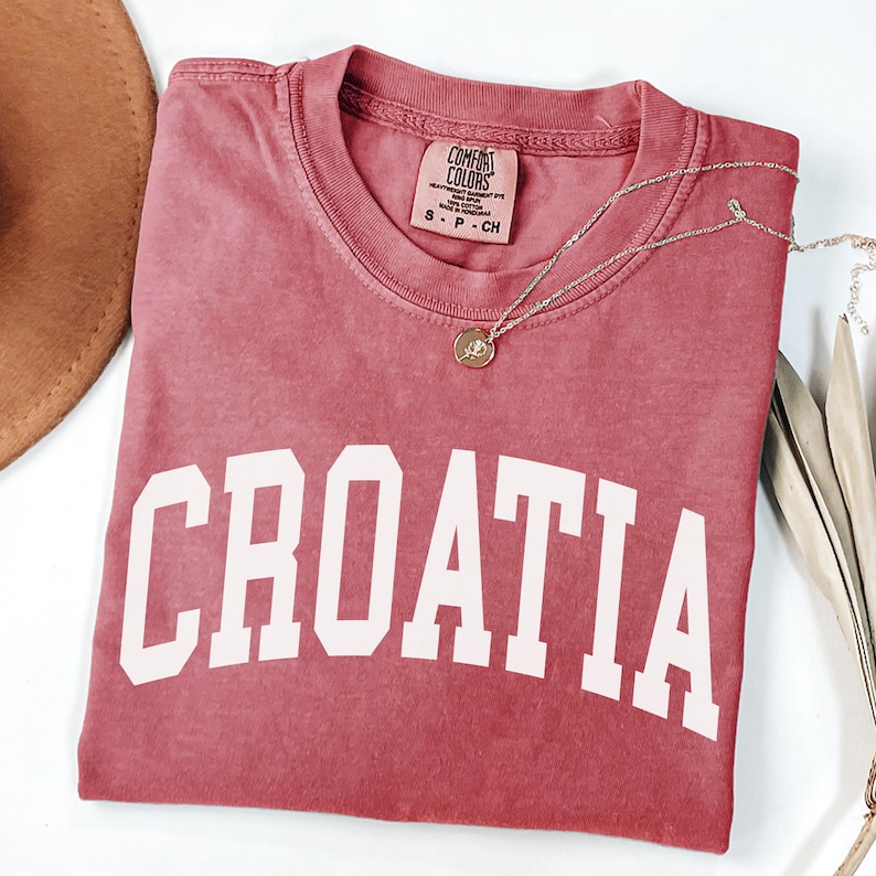 May include: A dusty rose-colored short-sleeve t-shirt featuring the word CROATIA printed in large white block letters across the chest. The shirt has a crew neckline and a Comfort Colors brand label inside the collar, along with a gold necklace.