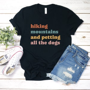 May include: Black t-shirt with the text "hiking mountains and petting all the dogs" in colorful retro font. The shirt is laid out on a white wooden surface with a pair of white sneakers and blue denim shorts.