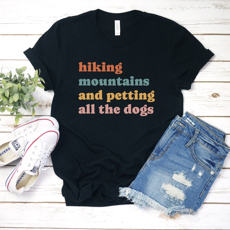 May include: Black t-shirt with the text "hiking mountains and petting all the dogs" in colorful retro font. The shirt is laid out on a white wooden surface with a pair of white sneakers and blue denim shorts.