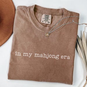 May include: A brown Comfort Colors t-shirt with the phrase "in my mahjong era" in white lettering. A silver necklace with a small pendant rests on the shirt. A brown hat is visible in the upper left corner.