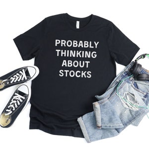 Puede incluir: Camiseta negra con texto blanco que dice "Probably Thinking About Stocks". La camiseta est&aacute; colocada sobre un fondo blanco con un par de zapatillas negras, unos vaqueros azules y unos auriculares verdes.