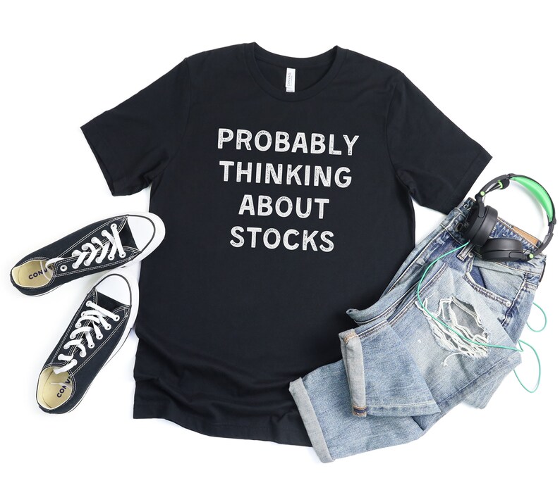 Puede incluir: Camiseta negra con texto blanco que dice "Probably Thinking About Stocks". La camiseta est&aacute; colocada sobre un fondo blanco con un par de zapatillas negras, unos vaqueros azules y unos auriculares verdes.