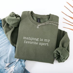 Puede incluir: Una sudadera de cuello redondo color verde oliva con el texto bordado en blanco "mahjong is my favorite sport" en el pecho. La prenda tiene mangas largas con pu&ntilde;os acanalados y un forro interior de forro polar suave, junto a un collar de plata.