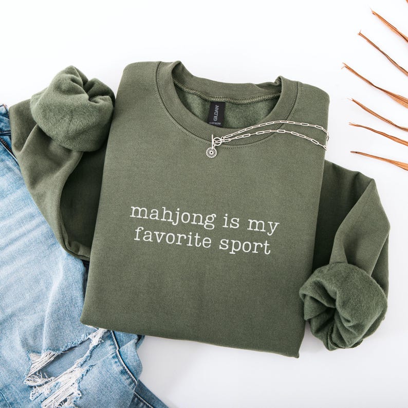 Puede incluir: Una sudadera de cuello redondo color verde oliva con el texto bordado en blanco "mahjong is my favorite sport" en el pecho. La prenda tiene mangas largas con pu&ntilde;os acanalados y un forro interior de forro polar suave, junto a un collar de plata.