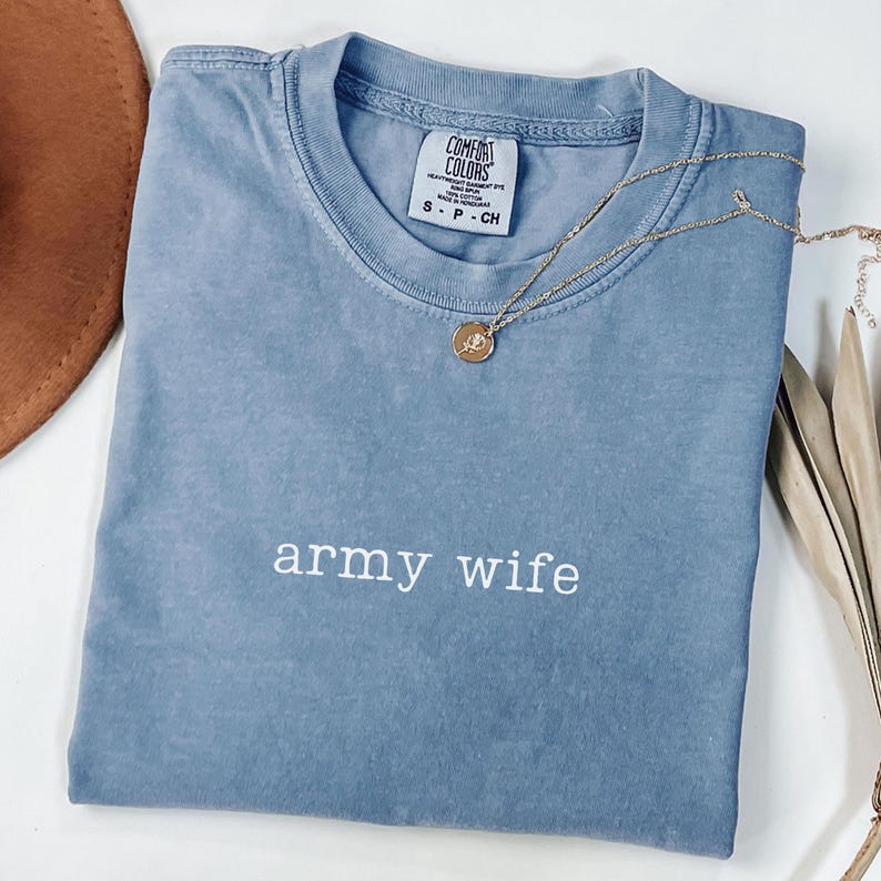 May include: A blue Comfort Colors t-shirt with the words "army wife" in white lettering. A gold necklace with a pendant rests on the shirt. The shirt is folded and placed on a white surface.