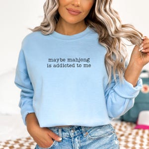 May include: A light blue sweatshirt with the text "maybe mahjong is addicted to me" in black. The sweatshirt is made of a soft material and is being worn by a person.