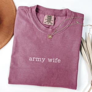 May include: A mauve Comfort Colors t-shirt with the words "army wife" in white. A gold-colored necklace with a pendant rests on the shirt. The shirt is folded and has a tag at the neckline. A brown hat is partially visible in the upper left corner.