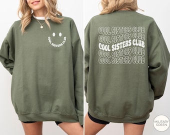 May include: A military green crewneck sweatshirt featuring a small white smiley face graphic with the text COOL SISTERS CLUB on the front left chest and a large, repeating wavy white text design reading COOL SISTERS CLUB on the back.