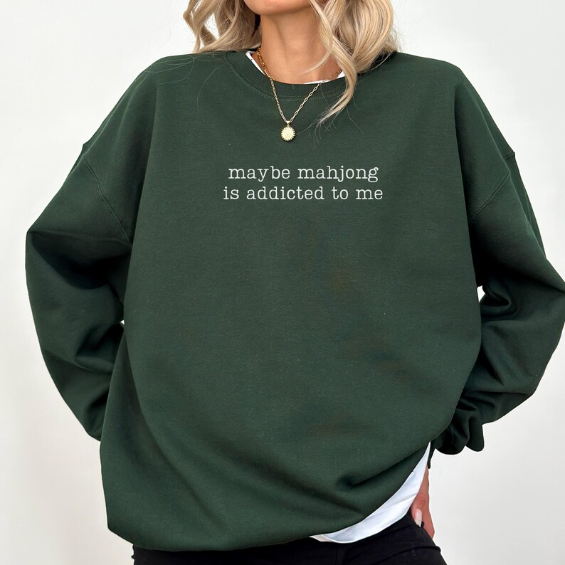 May include: Dark green sweatshirt with the text "maybe mahjong is addicted to me" in white. The sweatshirt has long sleeves and a crew neck. The model is wearing a gold necklace and a white undershirt.