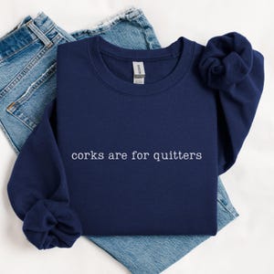 May include: A navy blue crewneck sweatshirt featuring the white embroidered text "corks are for quitters" centered on the chest. The garment has long sleeves with ribbed cuffs and a standard neckline, displayed over a pair of blue denim jeans.