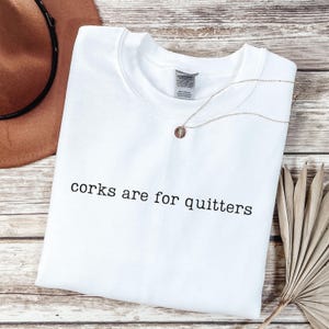 May include: A white crewneck sweatshirt featuring the black text "corks are for quitters" printed across the chest. The garment has a standard ribbed neckline and a visible interior brand label. A delicate gold-tone necklace rests on the fabric.