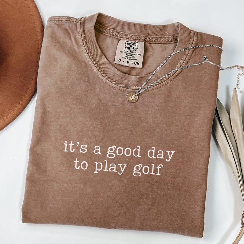 May include: A brown t-shirt with the text "it's a good day to play golf" in white. The shirt is made of a soft, comfortable material and has a relaxed fit.