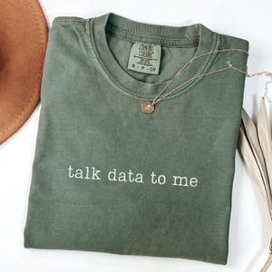 May include: An olive green t-shirt with the phrase "talk data to me" printed in white. A gold-colored necklace with a pendant is draped over the shirt. The shirt is folded neatly on a white surface.