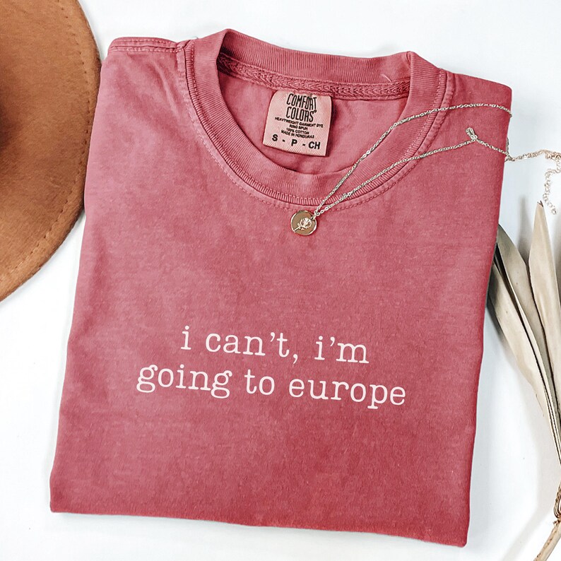 May include: A dusty rose short-sleeved t-shirt with a crew neckline and the white text "i can't, i'm going to europe" printed on the front. The shirt features a Comfort Colors brand label at the inner neck and is made of 100% cotton fabric.