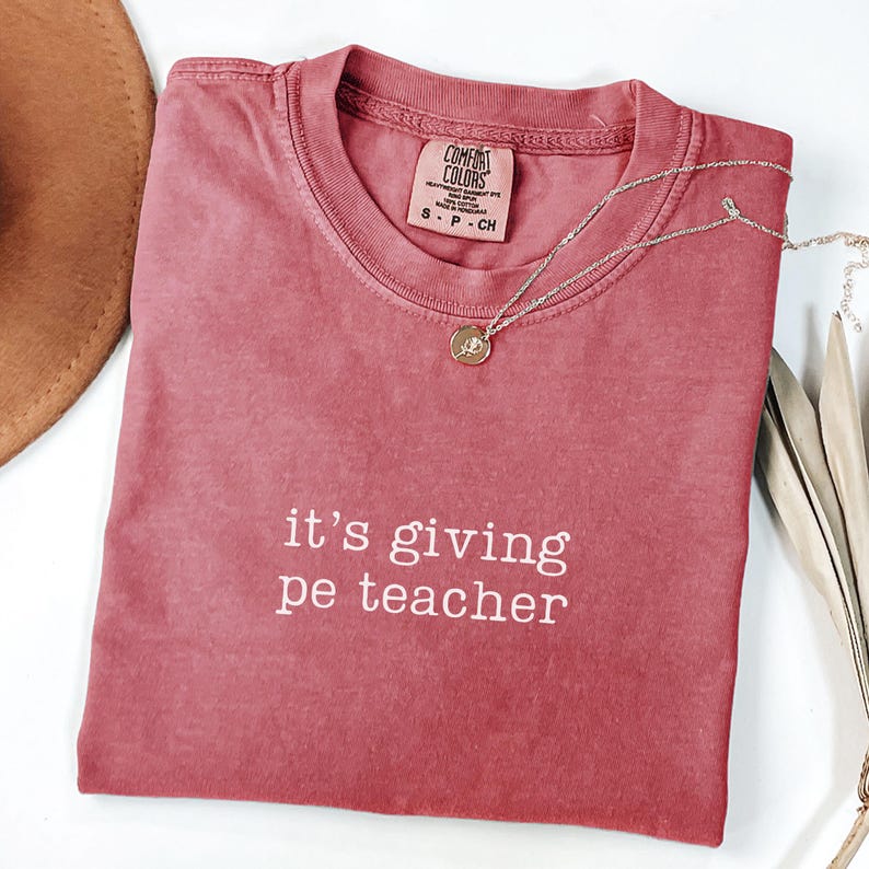 May include: A dusty rose Comfort Colors t-shirt with the phrase "it's giving pe teacher" in white lettering. A silver necklace with a small pendant rests on the shirt. The shirt is folded, and a brown hat is partially visible.