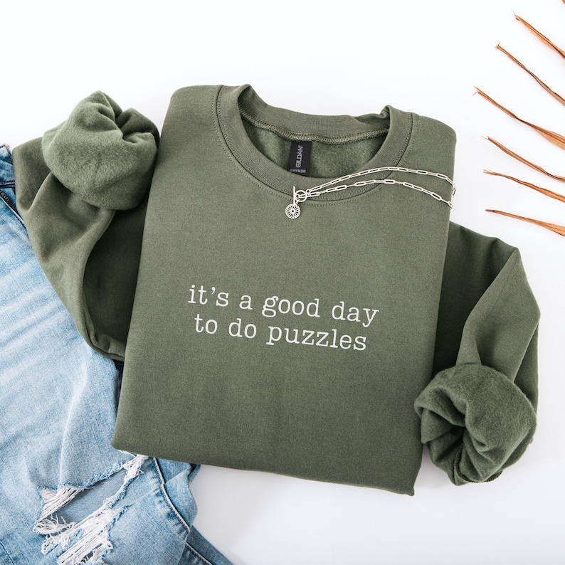 May include: An olive green crewneck sweatshirt featuring the white embroidered text "it's a good day to do puzzles" centered on the chest. The garment has long sleeves with ribbed cuffs and a silver-toned chain necklace resting on the neckline.