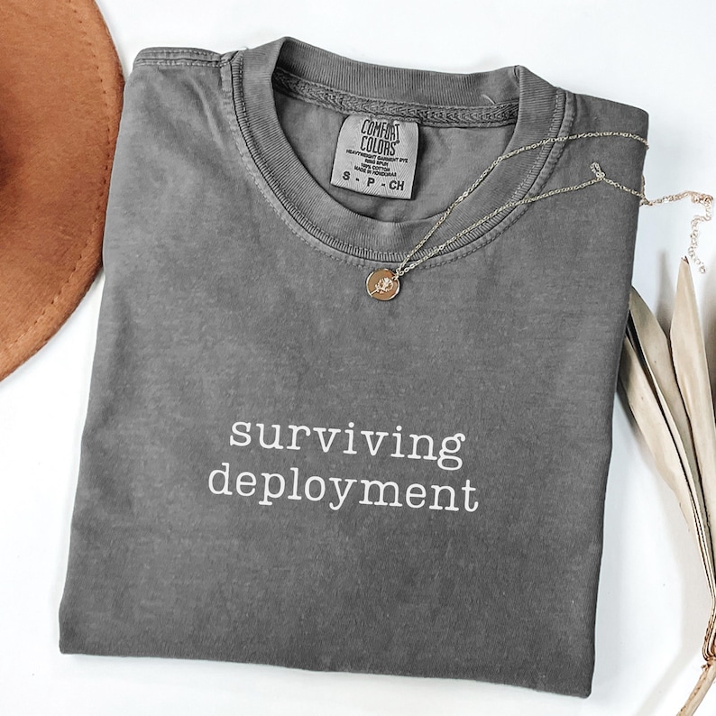 May include: A gray Comfort Colors t-shirt with the words "surviving deployment" in white. A silver necklace with a small pendant rests on the shirt. A brown hat and dried leaves are in the background.