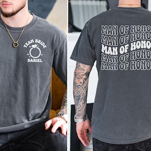 Pode incluir: Camiseta cinza carv&atilde;o com texto branco. A frente diz "TEAM BRIDE" acima de um gr&aacute;fico de anel e "DANIEL" abaixo. A parte de tr&aacute;s da camisa repete "MAN OF HONOR" em um design ondulado. Uma camiseta casual para um casamento.