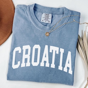 May include: A blue short-sleeve t-shirt with the word CROATIA printed in large white capital letters across the chest. The shirt features a Comfort Colors brand label at the inner neckline and is paired with a gold-toned circular pendant necklace.
