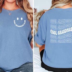 May include: A light blue t-shirt with the text "COOL GRANDMAS CLUB" on the back in white, stacked and wavy. The front features a smiley face design with the words "COOL GRANDMAS CLUB" in a semi-circle.