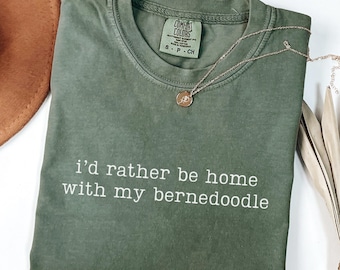 May include: An olive green t-shirt with the text "I'd rather be home with my bernedoodle" in white. A gold necklace with a pendant rests on the shirt. The shirt is folded and has a tag that says "Comfort Colors".