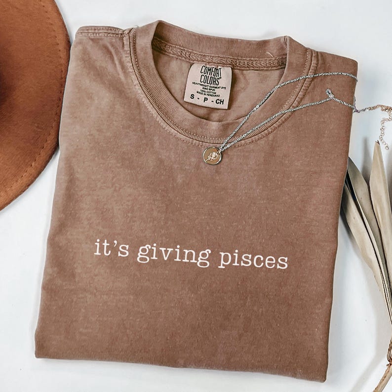 Puede incluir: Una camiseta marr&oacute;n Comfort Colors con la frase "it's giving pisces" en blanco. Un collar plateado con un peque&ntilde;o colgante est&aacute; sobre la camiseta. La camiseta est&aacute; doblada y se ve parcialmente un sombrero marr&oacute;n.