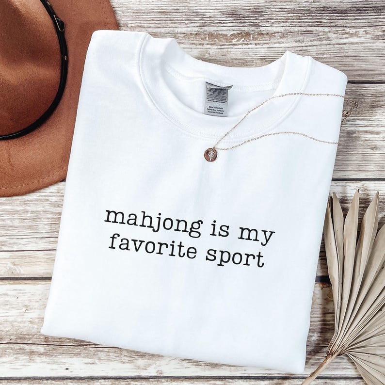 Puede incluir: Una sudadera blanca de cuello redondo con el texto negro "mahjong is my favorite sport" impreso en la parte delantera. La prenda tiene un cuello acanalado est&aacute;ndar y mangas largas, presentada como una pieza de ropa doblada con un dise&ntilde;o sencillo.
