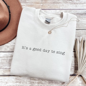 May include: A light beige sweatshirt with the phrase "it's a good day to sing" printed in black. A gold necklace with a pendant rests on the sweatshirt. A brown hat and dried plant are in the background.