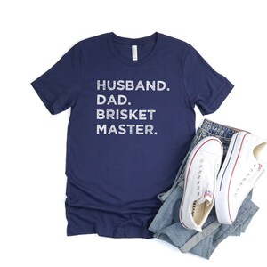 May include: Navy blue t-shirt with white text that reads "Husband. Dad. Brisket Master."