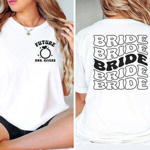 May include: White t-shirt with the text "FUTURE MRS. RIVERS" and a ring with a heart on the front. The back of the shirt has the word "BRIDE" repeated five times in a retro font.