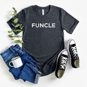 May include: A dark gray t-shirt with the word "FUNCLE" printed in white on the front. The shirt is laid flat on a white surface with a pair of black sneakers and a pair of blue jeans.