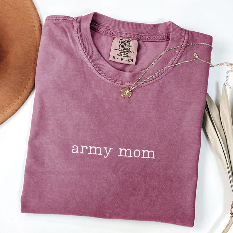 Puede incluir: Una camiseta Comfort Colors malva con las palabras "army mom" en escritura blanca. Un collar dorado con un colgante descansa sobre la camiseta. Un sombrero marr&oacute;n es visible en la esquina superior izquierda.