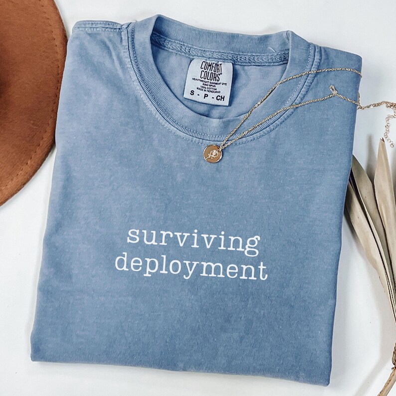 May include: A blue Comfort Colors t-shirt with the words "surviving deployment" in white. A gold necklace with a pendant rests on the shirt. The shirt is folded neatly.